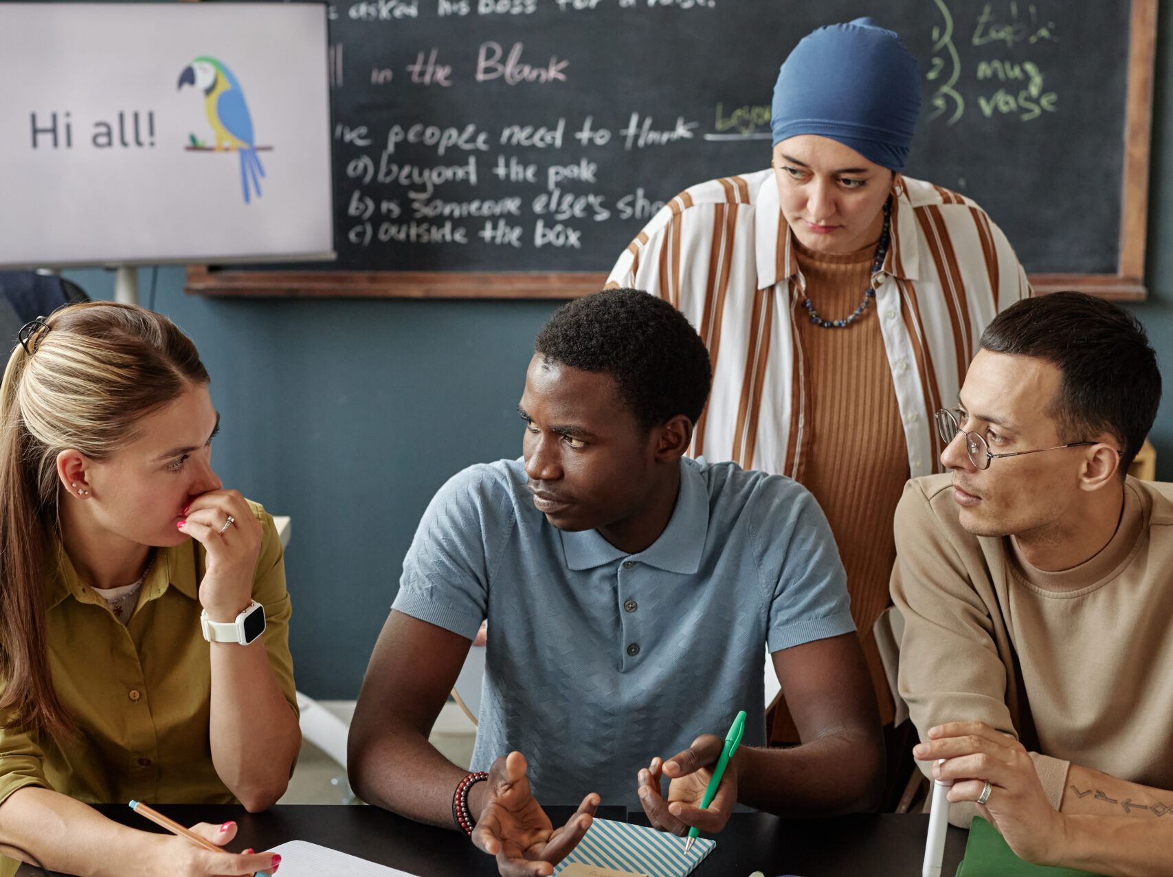Vertical shot of young Black man talking to female teacher sitting at desk in group discussion, while practicing new words in speaking club session at language school for adult learners